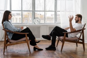psicóloga e paciente conversando frente a frente sentados em sala branca com janela grande e um relógio