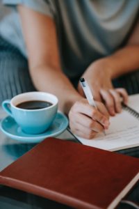pessoa escrevendo a caneta em um caderno com uma xícara de café ao lado