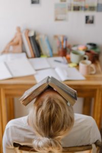 mulher jovem com livro na cara e cabeça para trás em frente a mesa cheia de papéis cansada da leitura diária